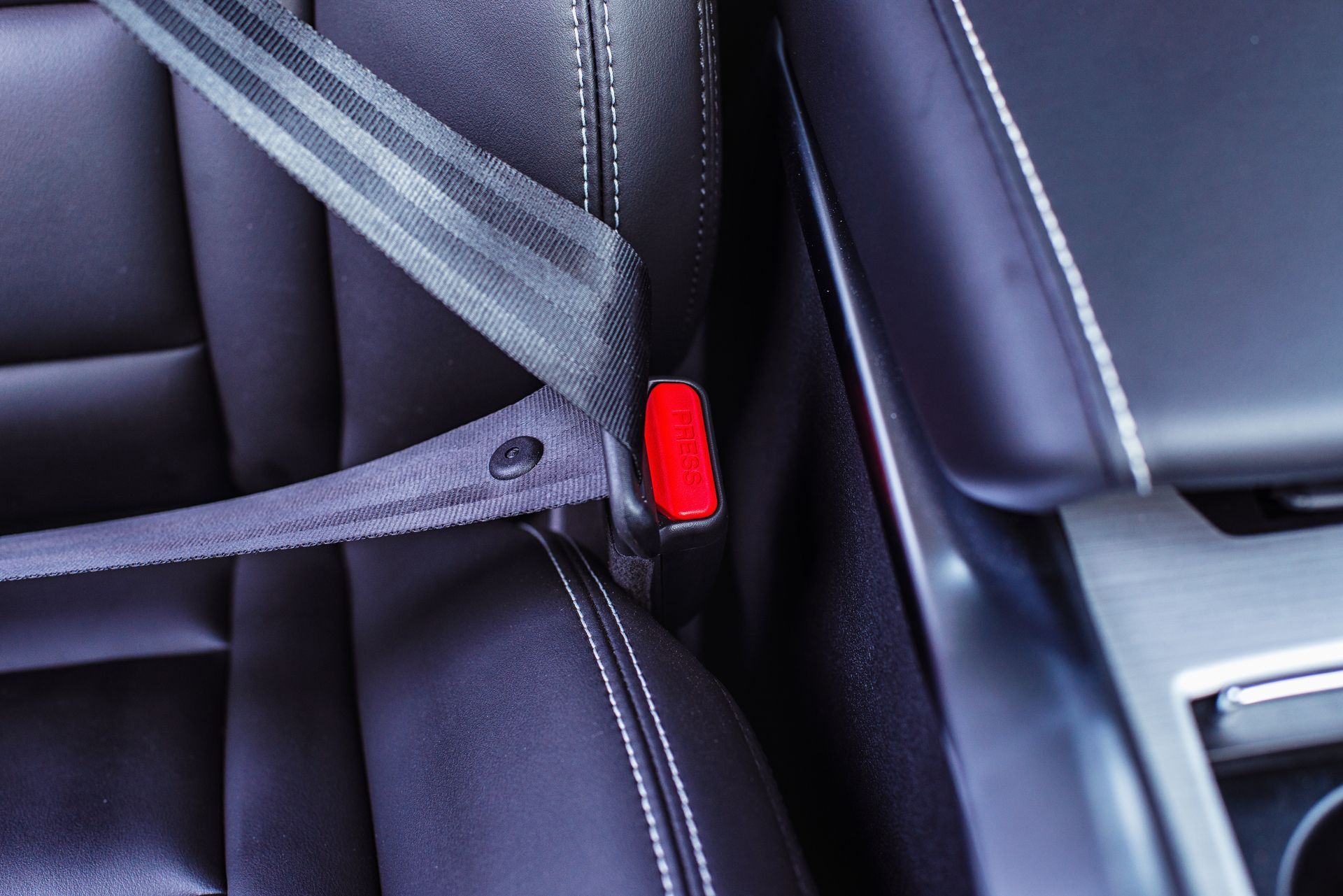 A dark leather car seat with a seatbelt fastened into a red-buttoned buckle.