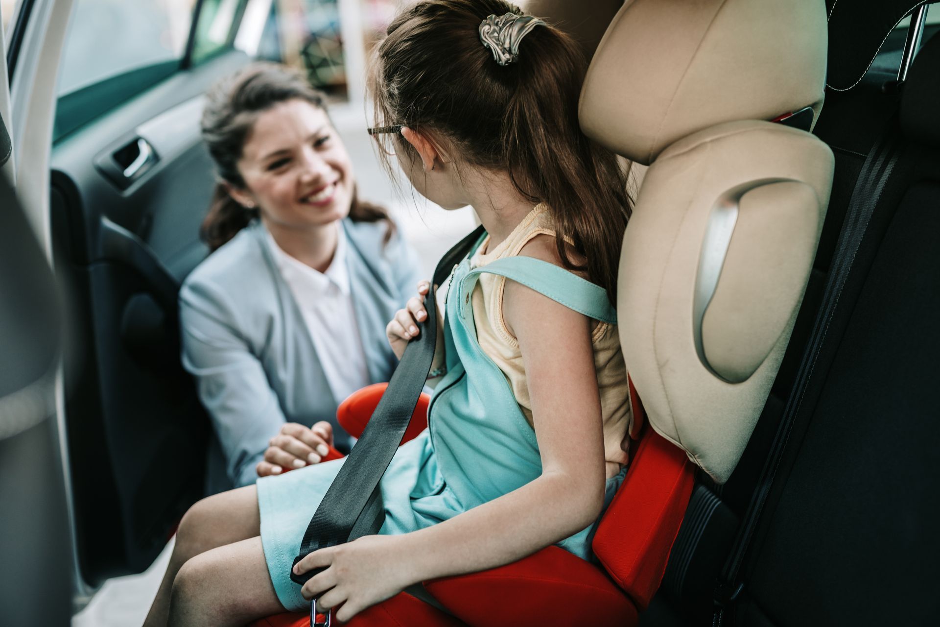 A person in a blazer helps a child fasten their seatbelt while sitting in a red booster seat in the back of a car.