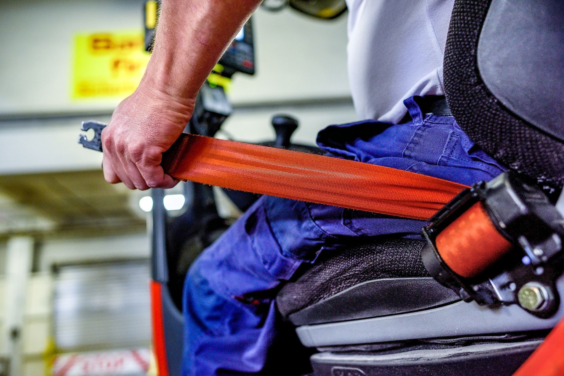 A close-up of a person wearing blue work pants fastening an orange safety seatbelt while seated in a vehicle.