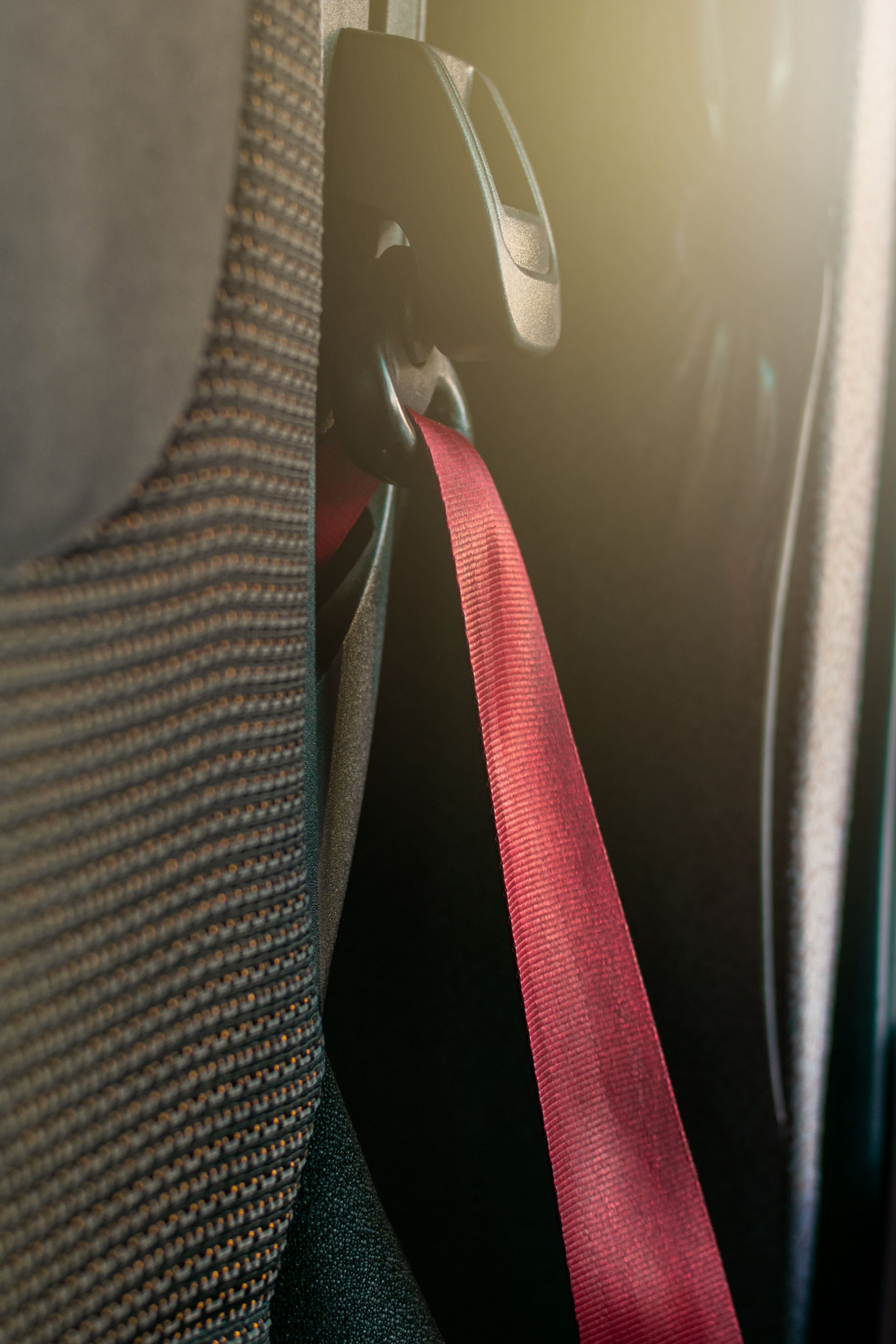 A red seatbelt strap hanging from a black retractor assembly mounted to the side of a textured grey car seat.