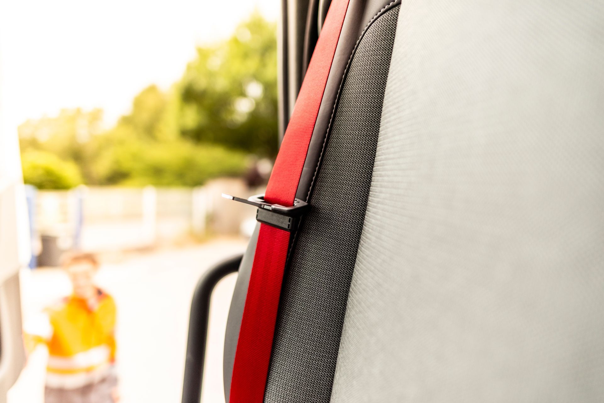 A close-up view of a red seatbelt strap against a gray seat, with a blurred outdoor background.
