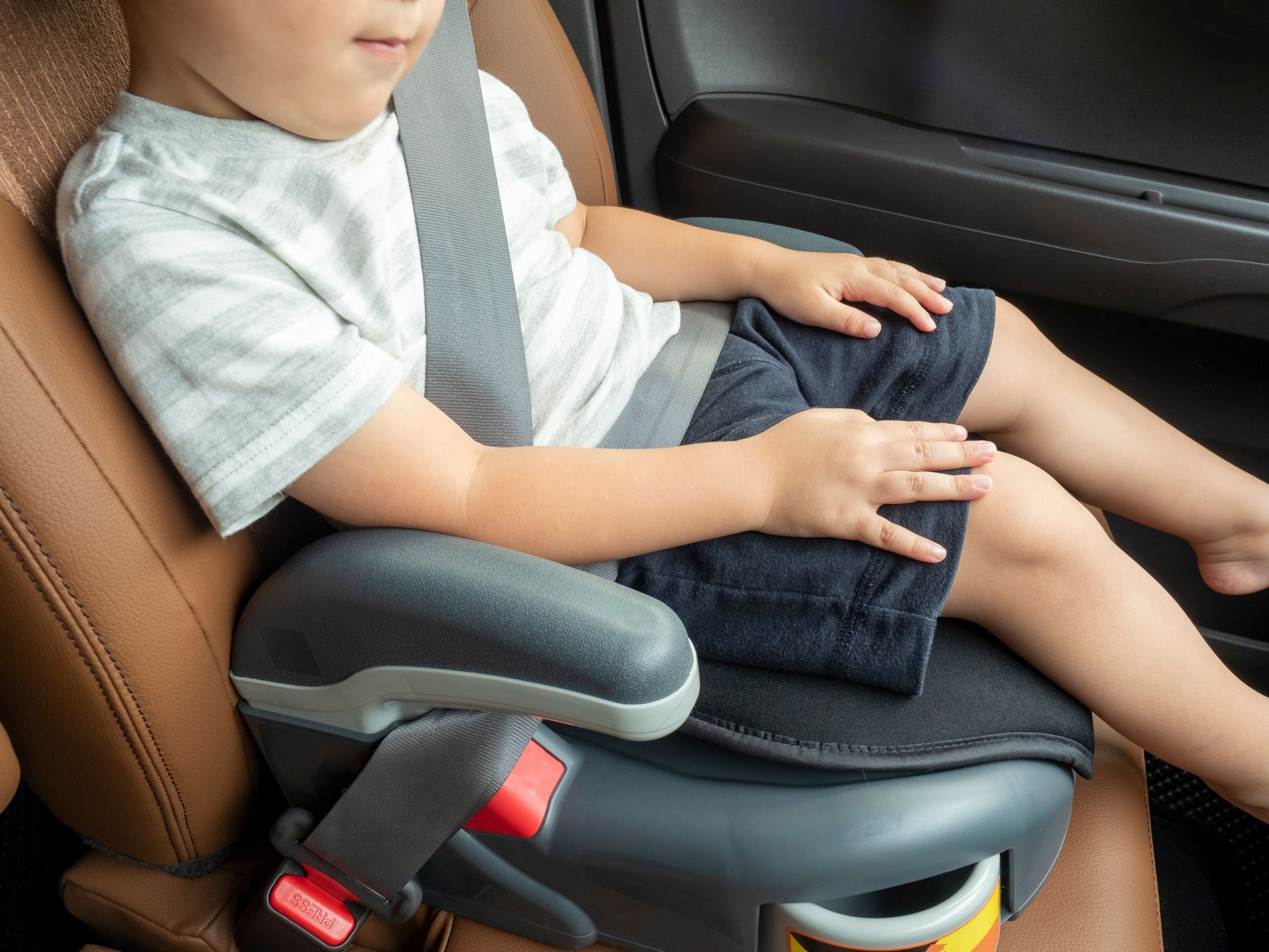 A child seated in a booster seat in a car, wearing a gray striped t-shirt and dark shorts, secured by a seat belt.