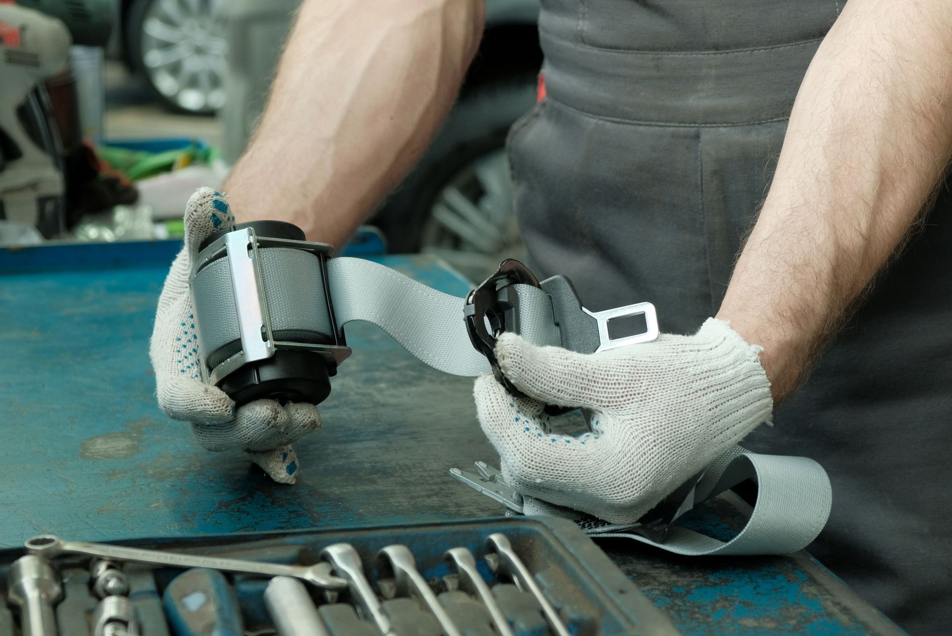 A mechanic wearing gloves inspects a vehicle seat belt mechanism on a workbench filled with tools.