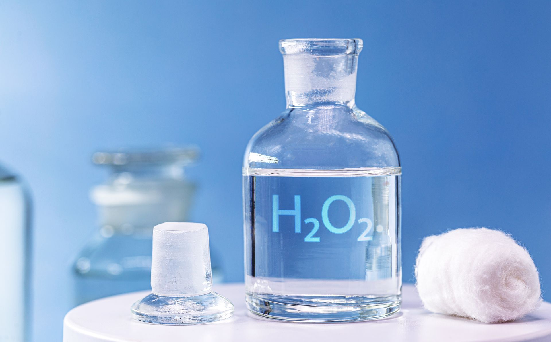 A glass bottle labeled H2O2 sits on a white surface next to its glass stopper and a small roll of white medical gauze.