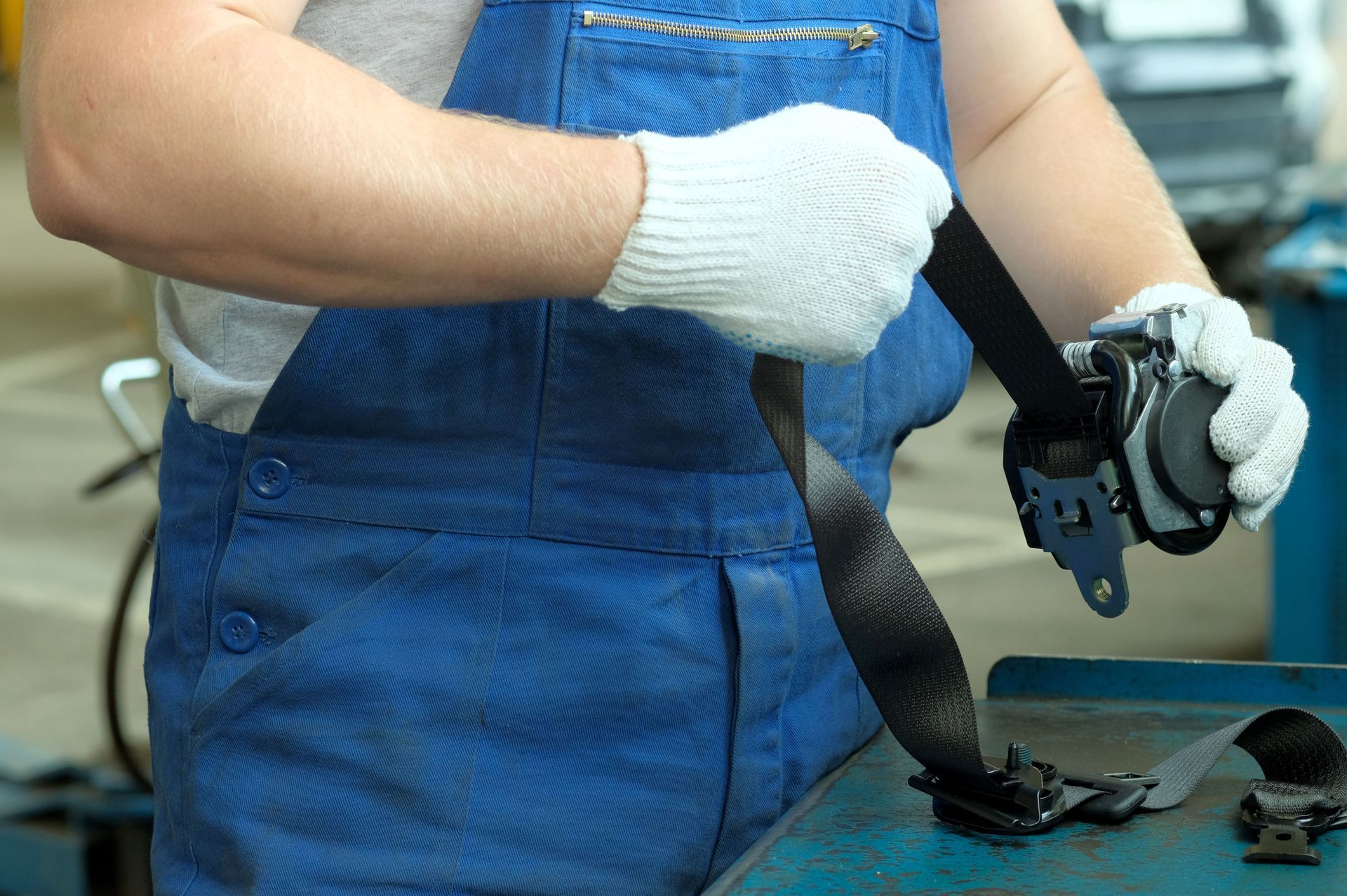 A person in blue work overalls and white gloves holds a black car seat belt retractor assembly.