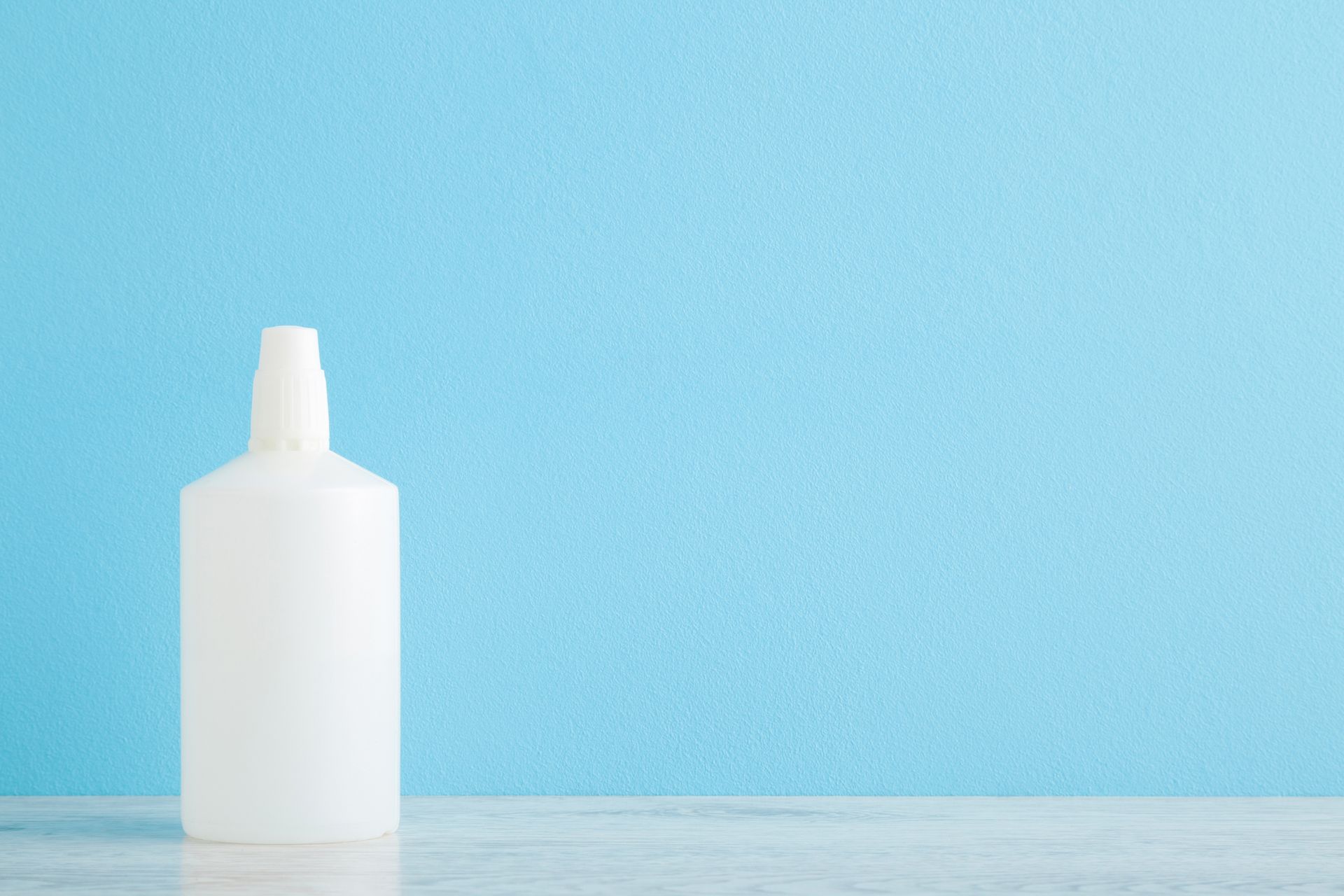 A white, opaque plastic bottle sits on a marble surface against a solid, light blue background.