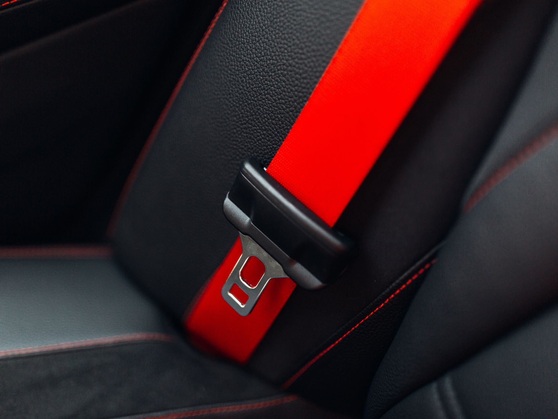 A bright red seatbelt with a black buckle against a black leather car seat with red stitching.