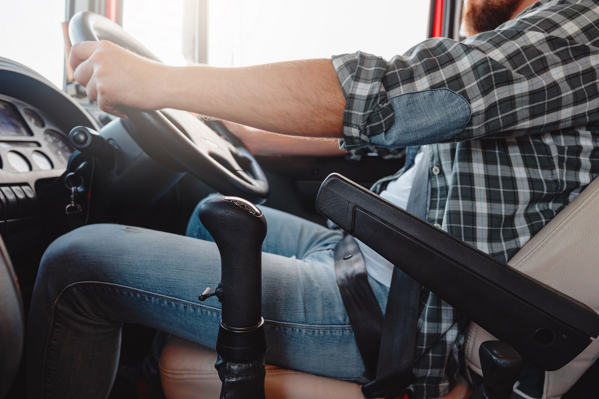 A driver in a plaid shirt and jeans grips the steering wheel of a vehicle with a prominent gear shift in the foreground.