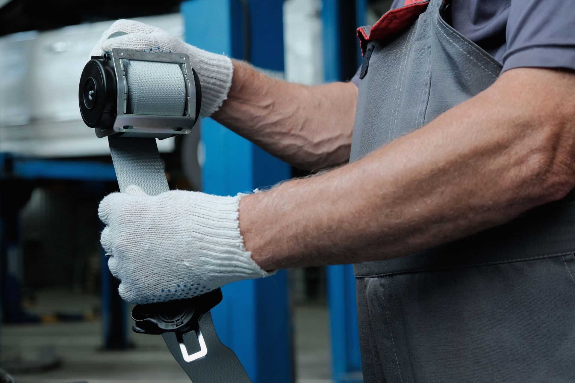 A mechanic in gray work clothes and white gloves holds a car seat belt tensioner assembly in a garage.