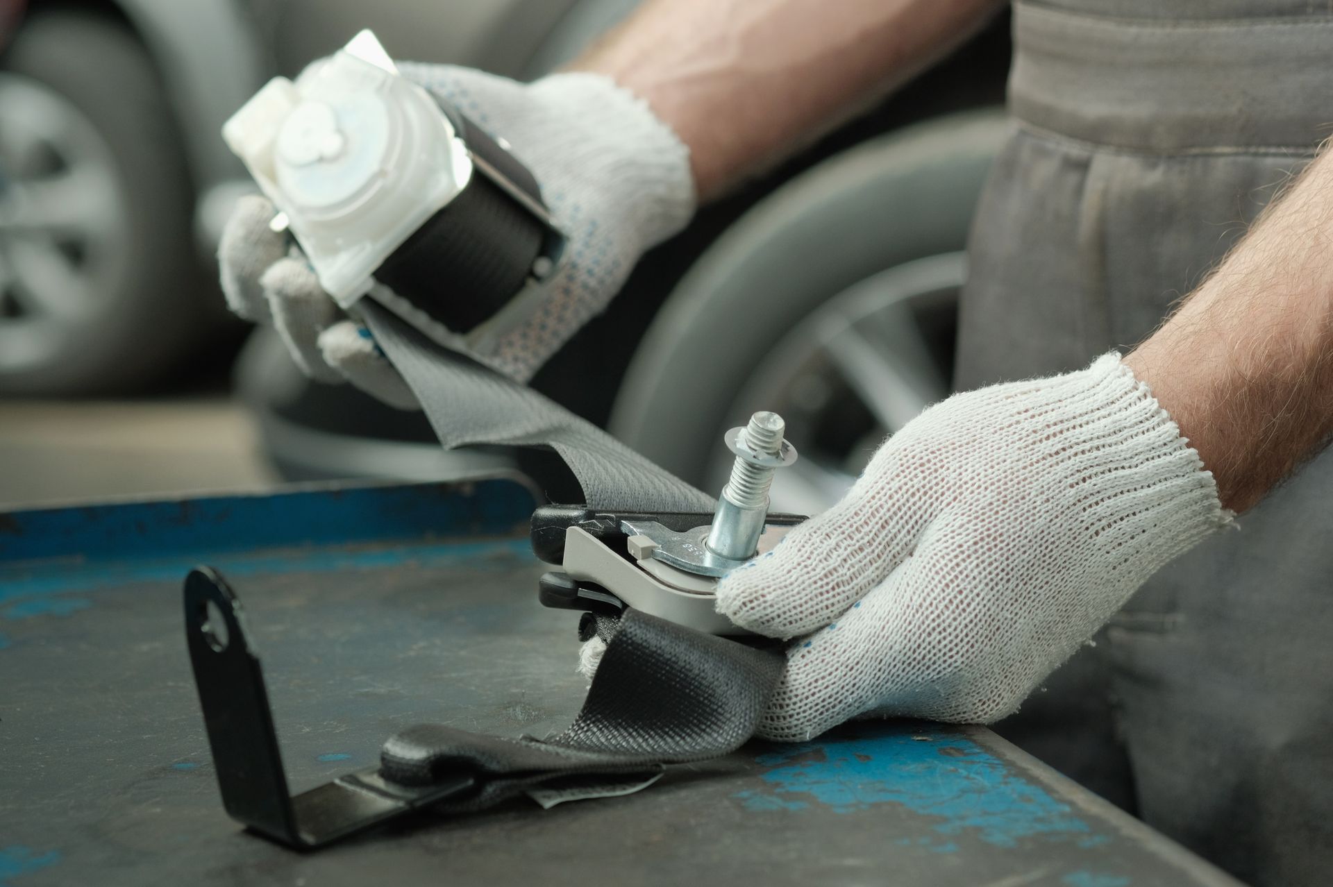 A technician wearing gloves works on a car seatbelt mechanism at a workbench in a repair shop.