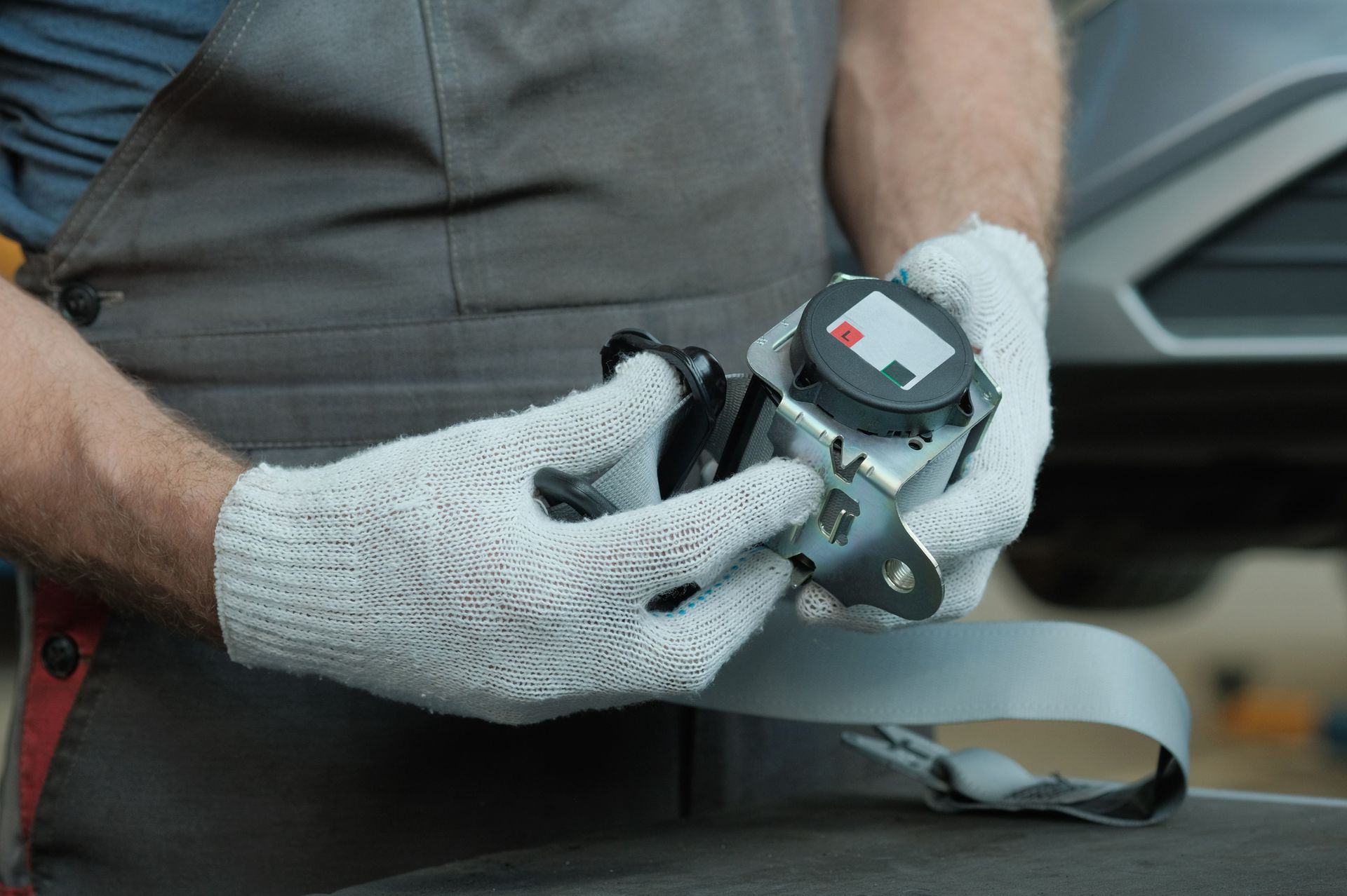 A mechanic wearing white gloves inspects a gray car seat belt retraction mechanism.