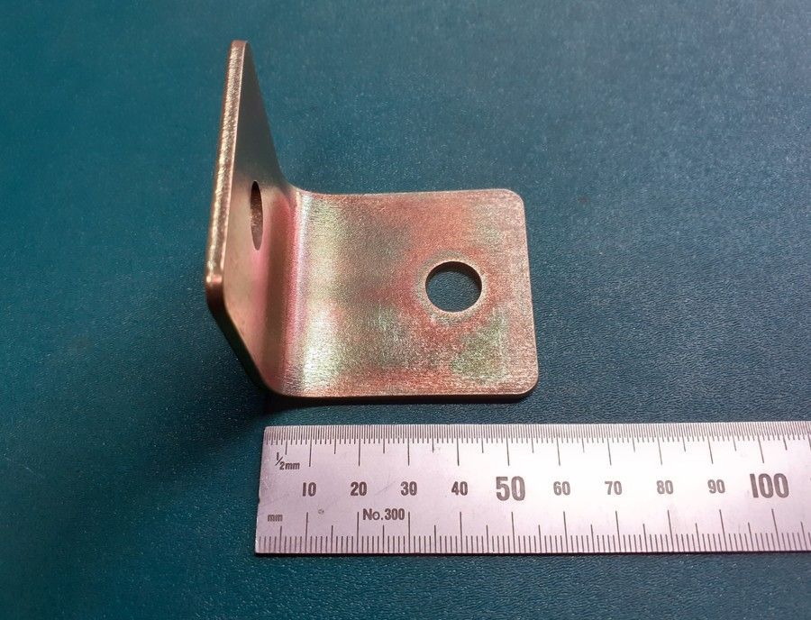 Metal L-bracket with a circular hole, shown beside a ruler on a blue background.