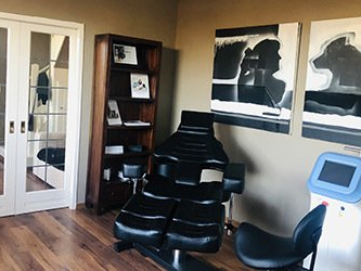 Een behandelkamer met een zwarte relaxfauteuil, een rolkruk, een hoge houten boekenkast en abstracte muurkunst.