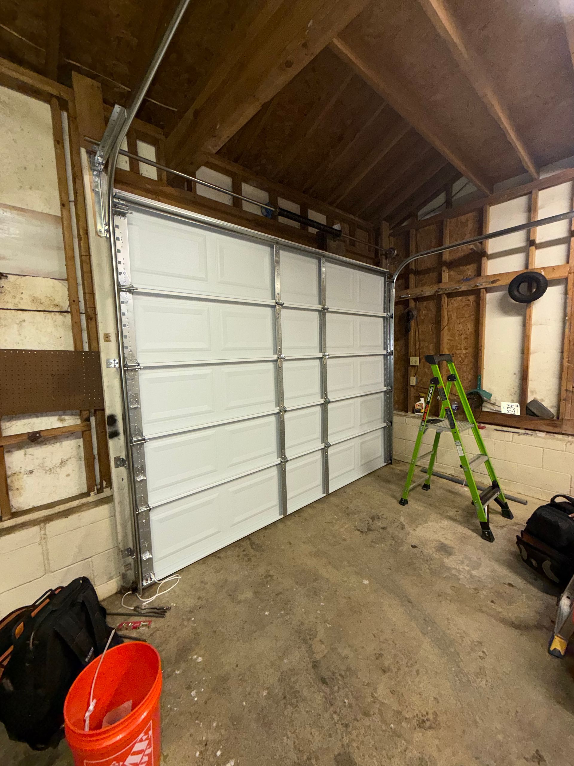 Interior of a garage with a white sectional door, concrete floor, ladder, and bags under a wooden ceiling.