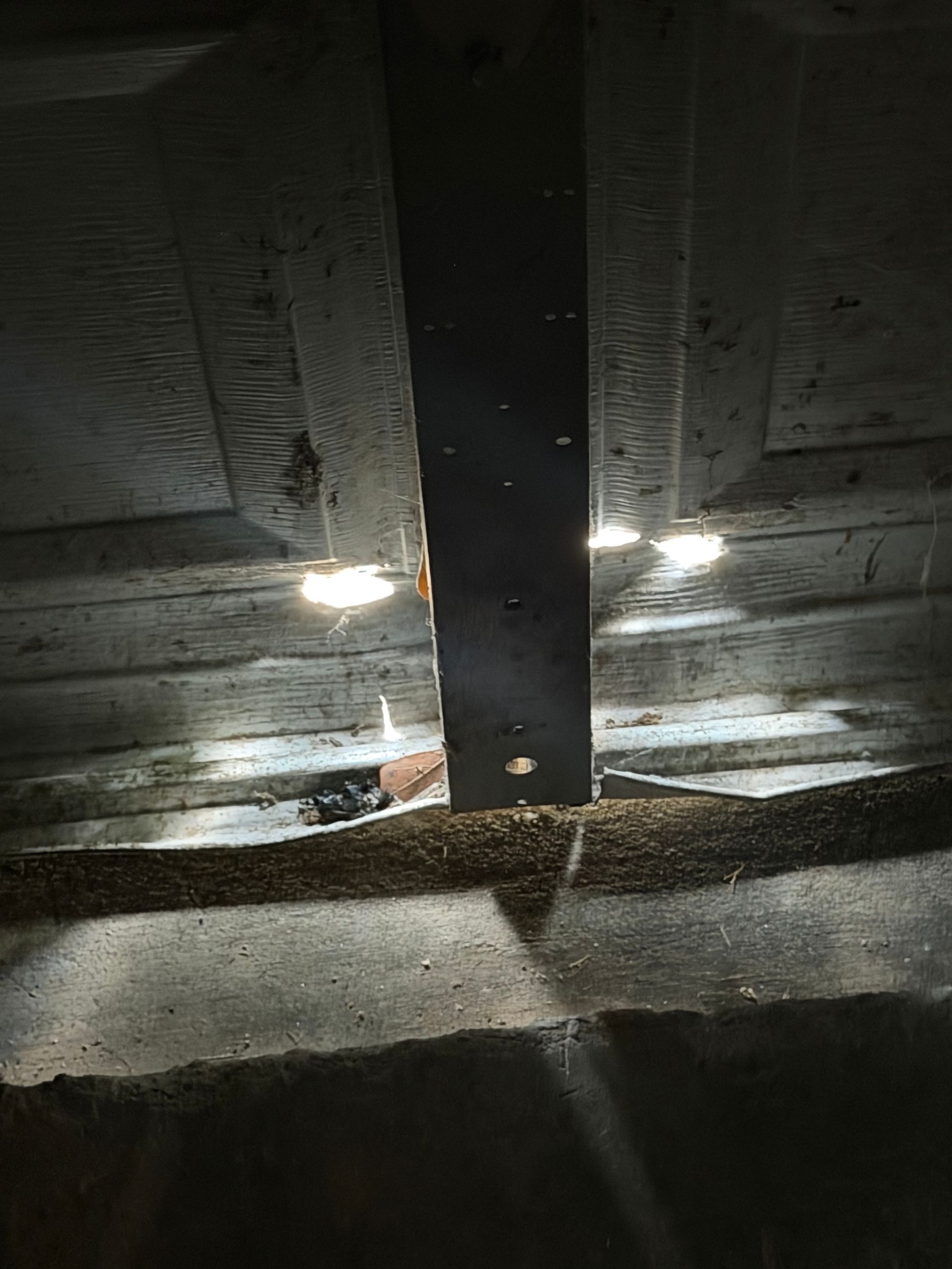 Dim attic ceiling with a black support beam and two bright lights shining on insulation sheets