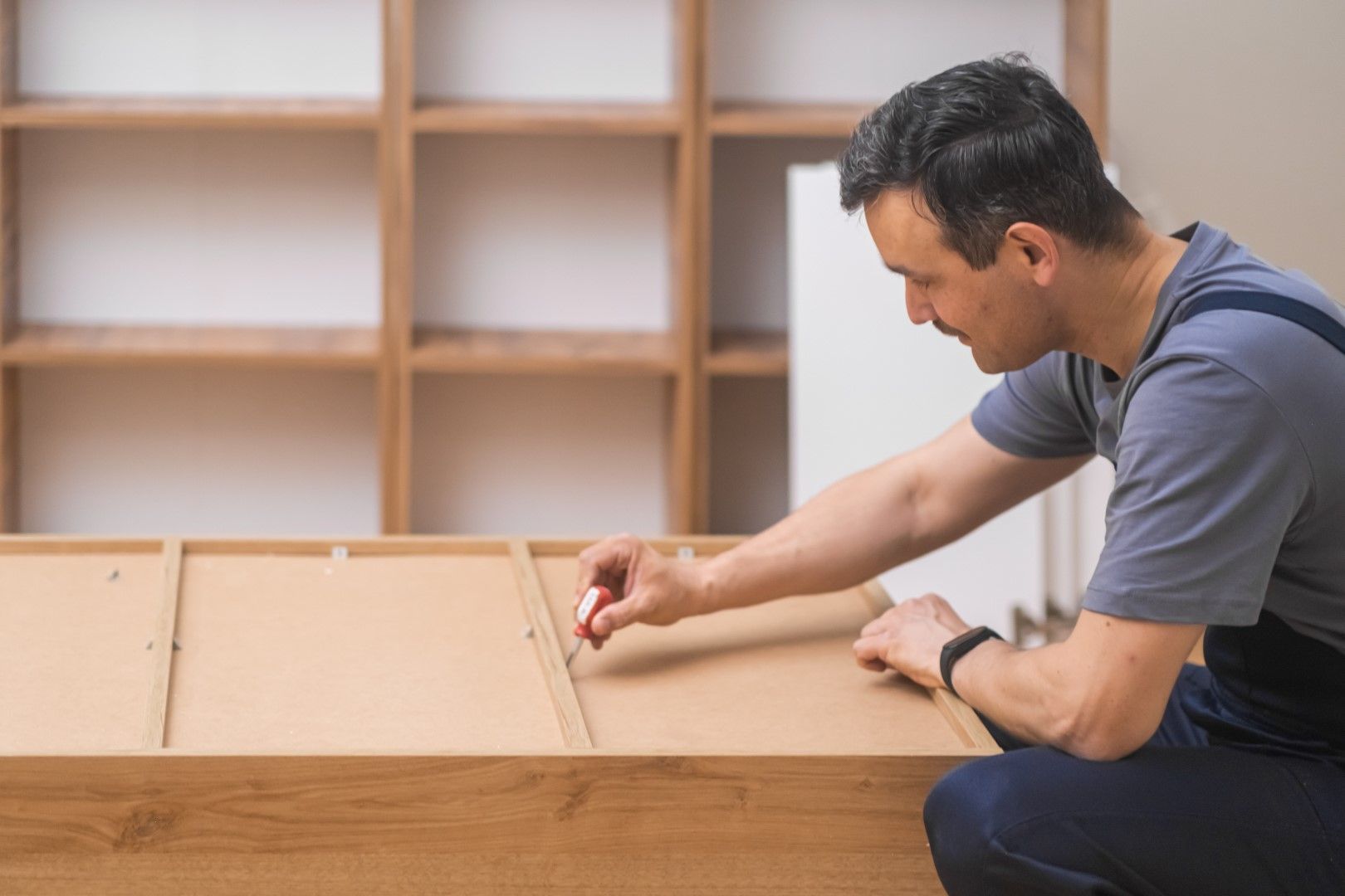 Man assembling furniture, using a tool, in a room with a wooden bookcase in the background.
