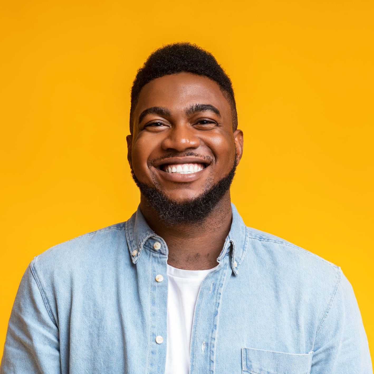 Smiling Black man with a beard wearing a light blue button-down shirt and white t-shirt, yellow background.