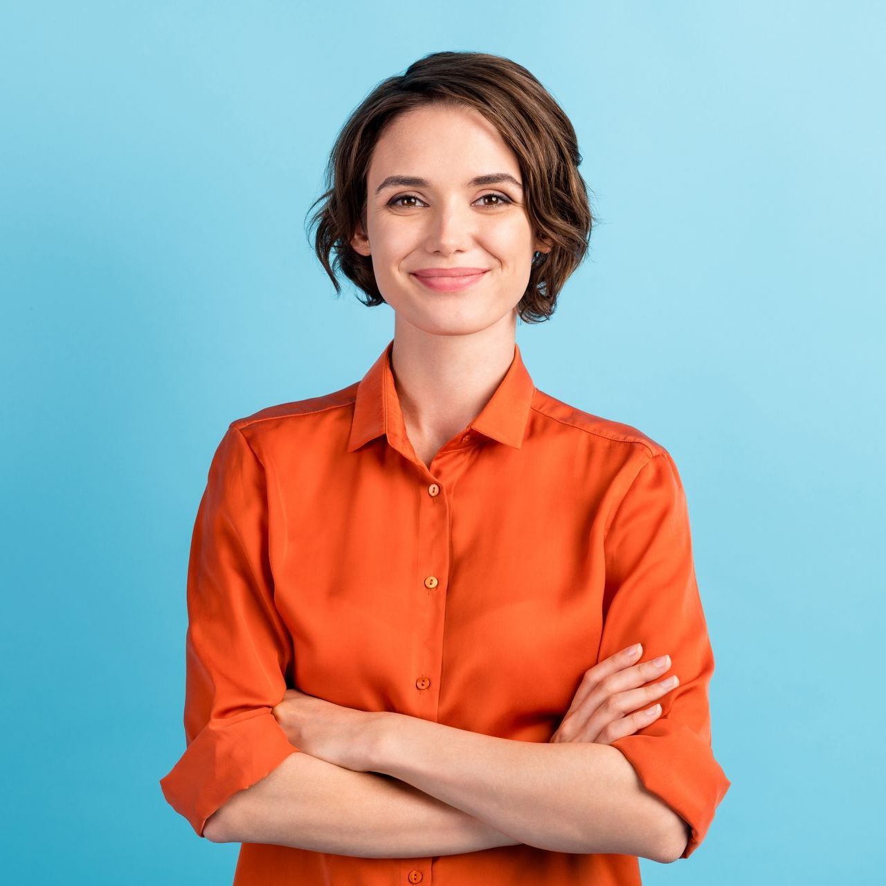 Woman with short brown hair, orange shirt, arms crossed, smiling against a blue backdrop.