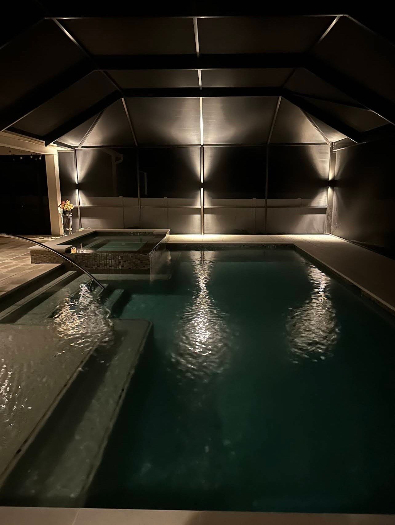 Pool at night with overhead lighting illuminating water, walls, and a hot tub.