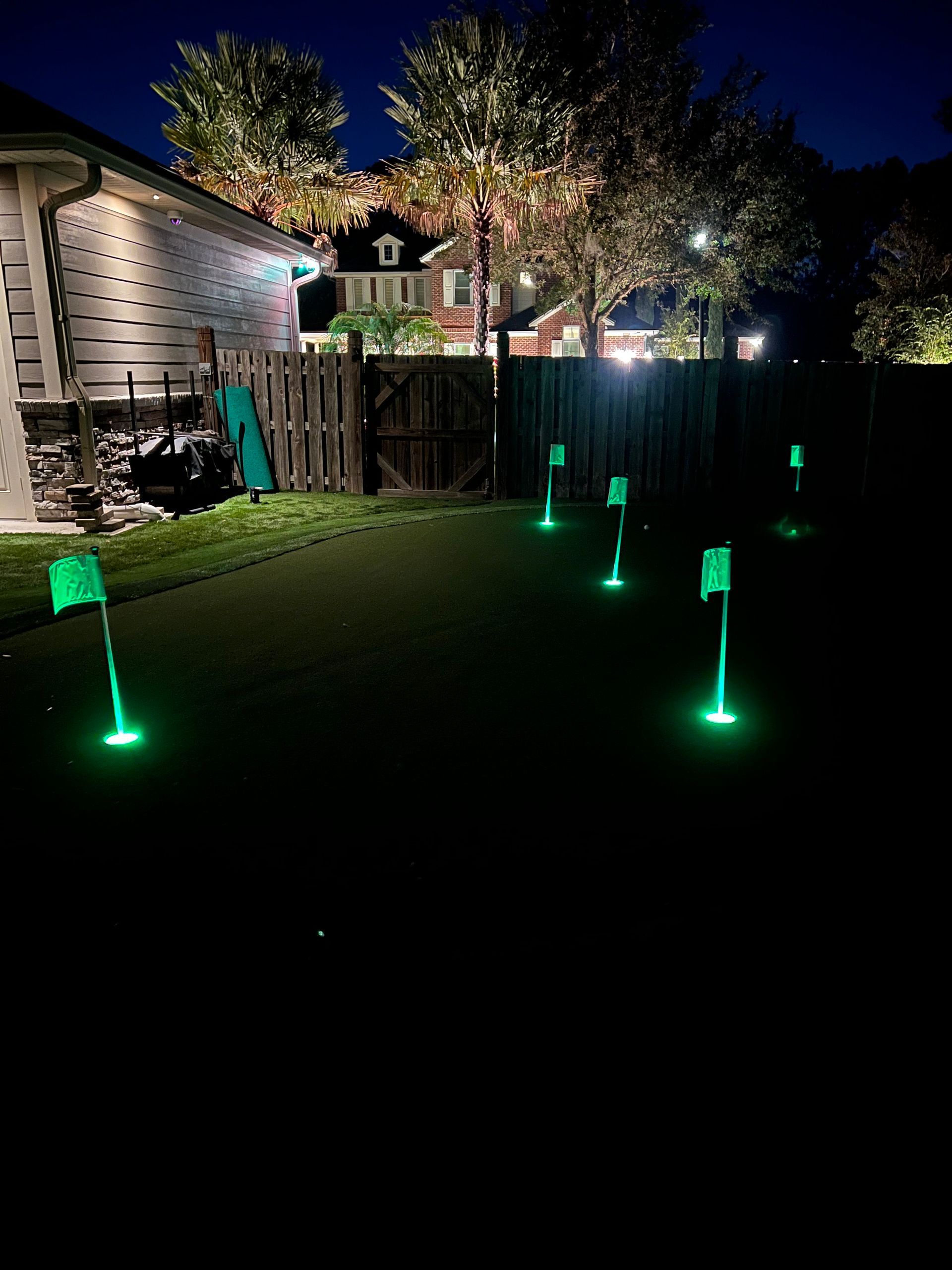 Backyard putting green at night, lit green flags, illuminated trees and houses in background.