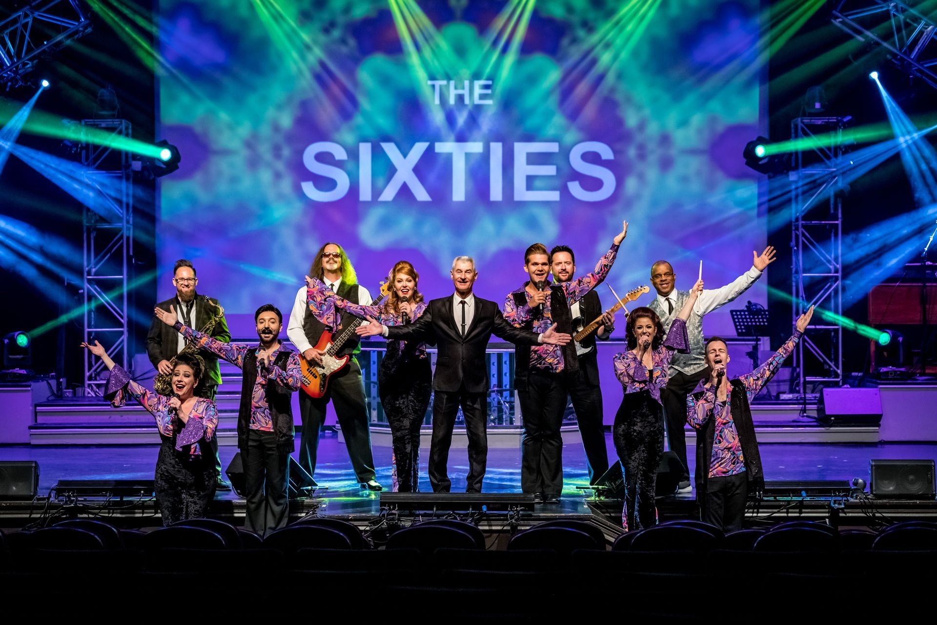 A band and performers pose on a brightly lit stage in front of a screen displaying 
