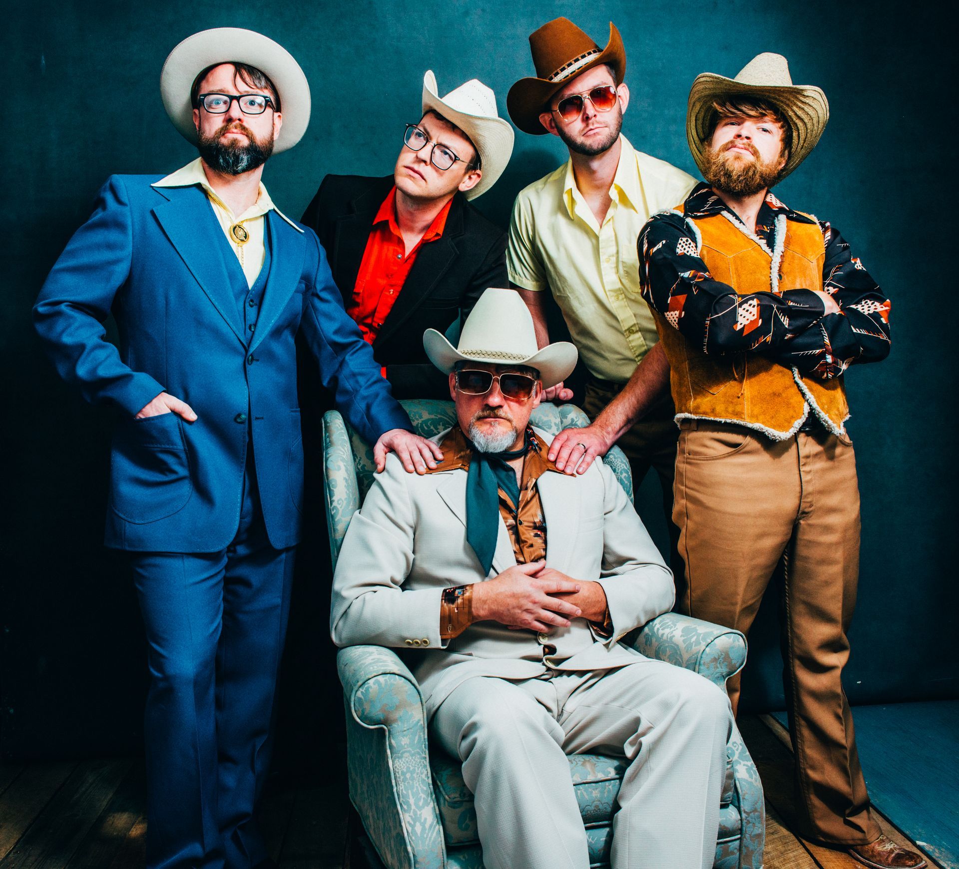 Five people in western-style suits and hats pose against a dark blue background; one sits in a chair, four stand behind.