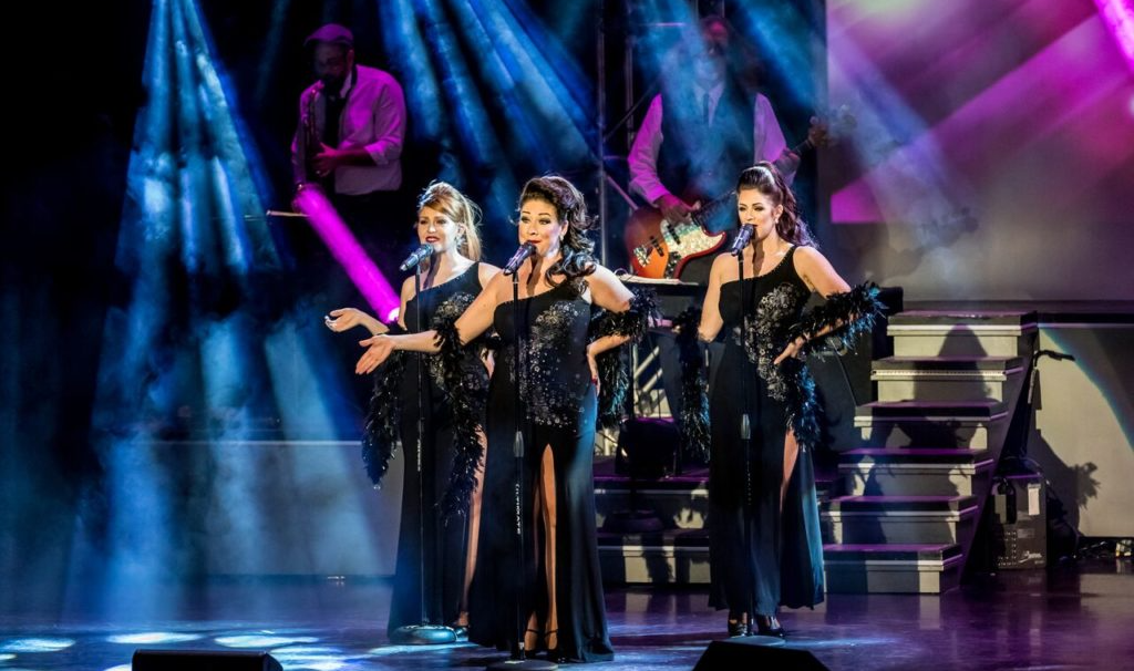 Three performers in black dresses sing on a stage with blue lighting, accompanied by a band in the background.