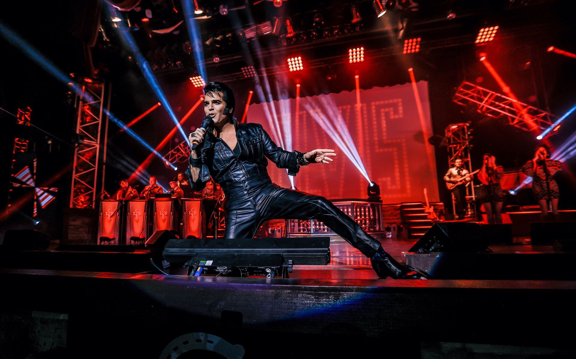 A performer in a black suit singing on stage with bright red lights, blue spotlights, and a background banner reading ELVIS.