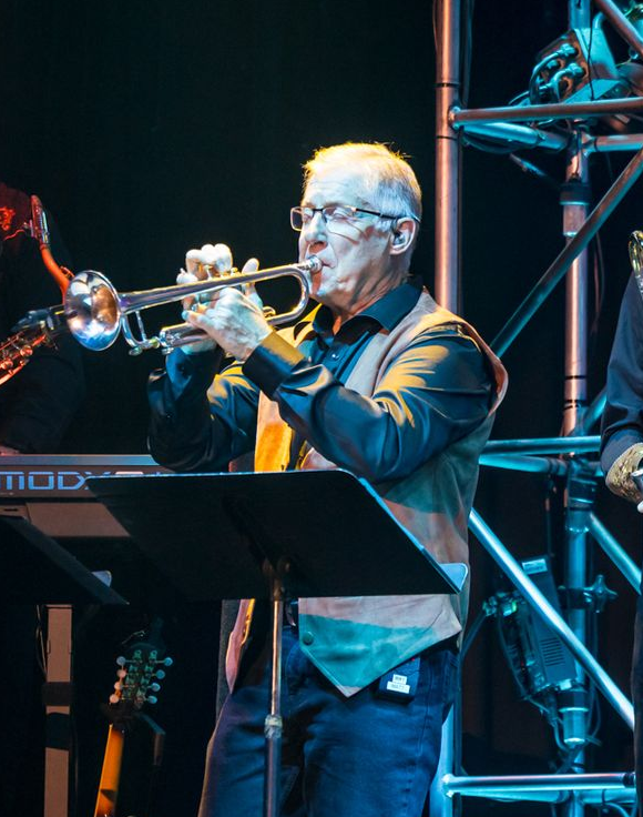 A musician in a black shirt and vest plays a trumpet on stage behind a music stand 