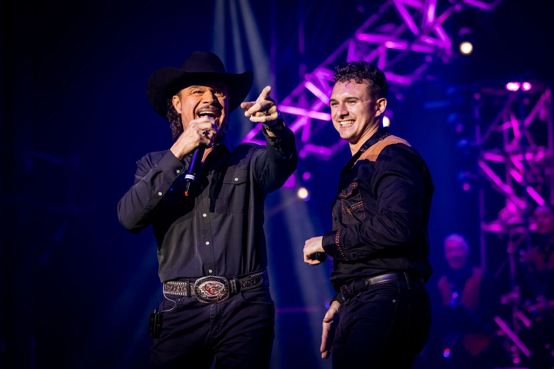 A performer in a cowboy hat and vest sings into a microphone on stage, with a guitar silhouette visible on the screen.