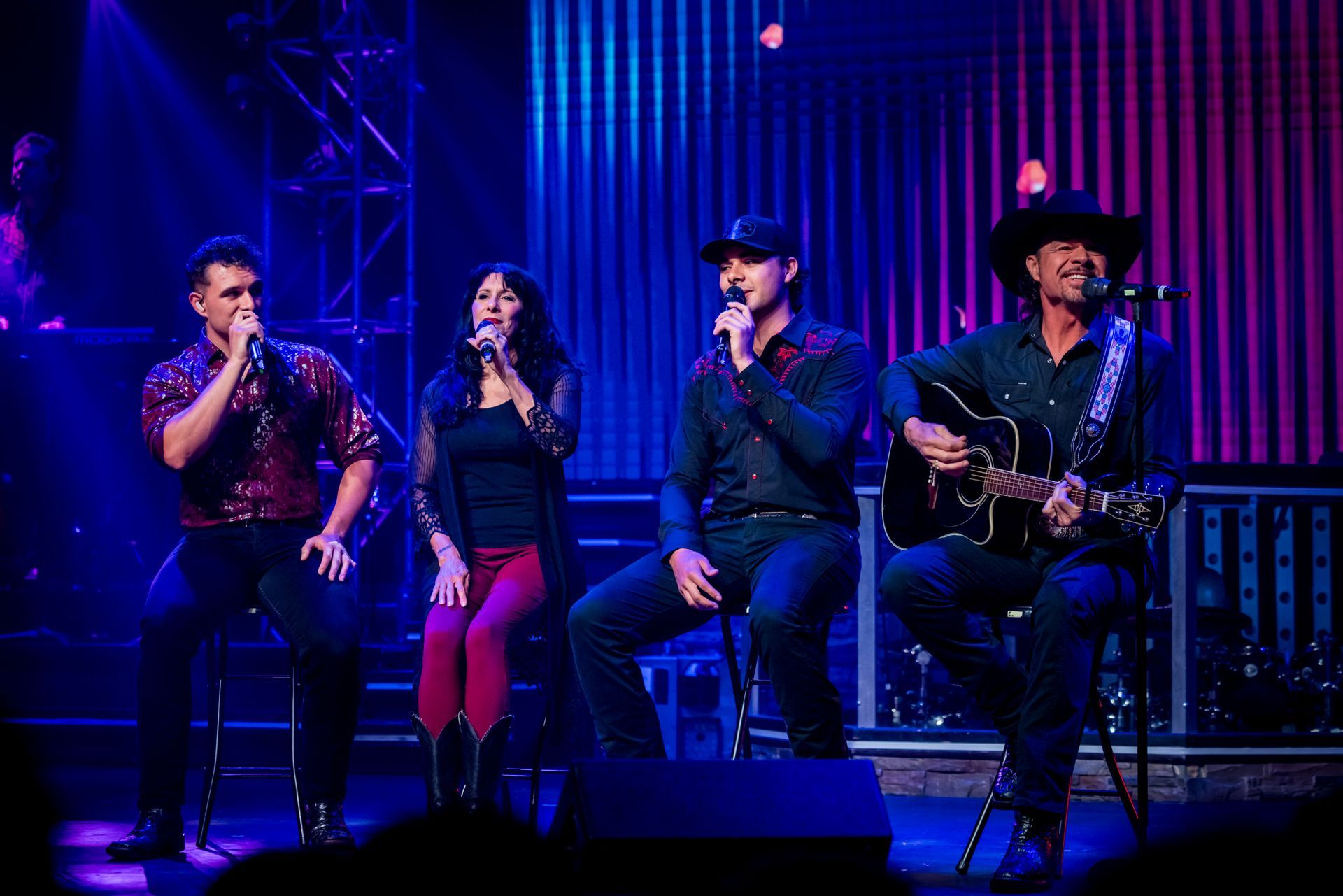 A man in a cowboy hat and vest singing into a microphone alongside a woman in a light dress on a brightly lit stage.