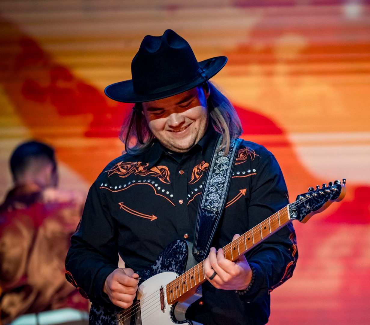 A musician wearing a cowboy hat and western-style shirt plays an electric guitar on a stage with a blue light background.