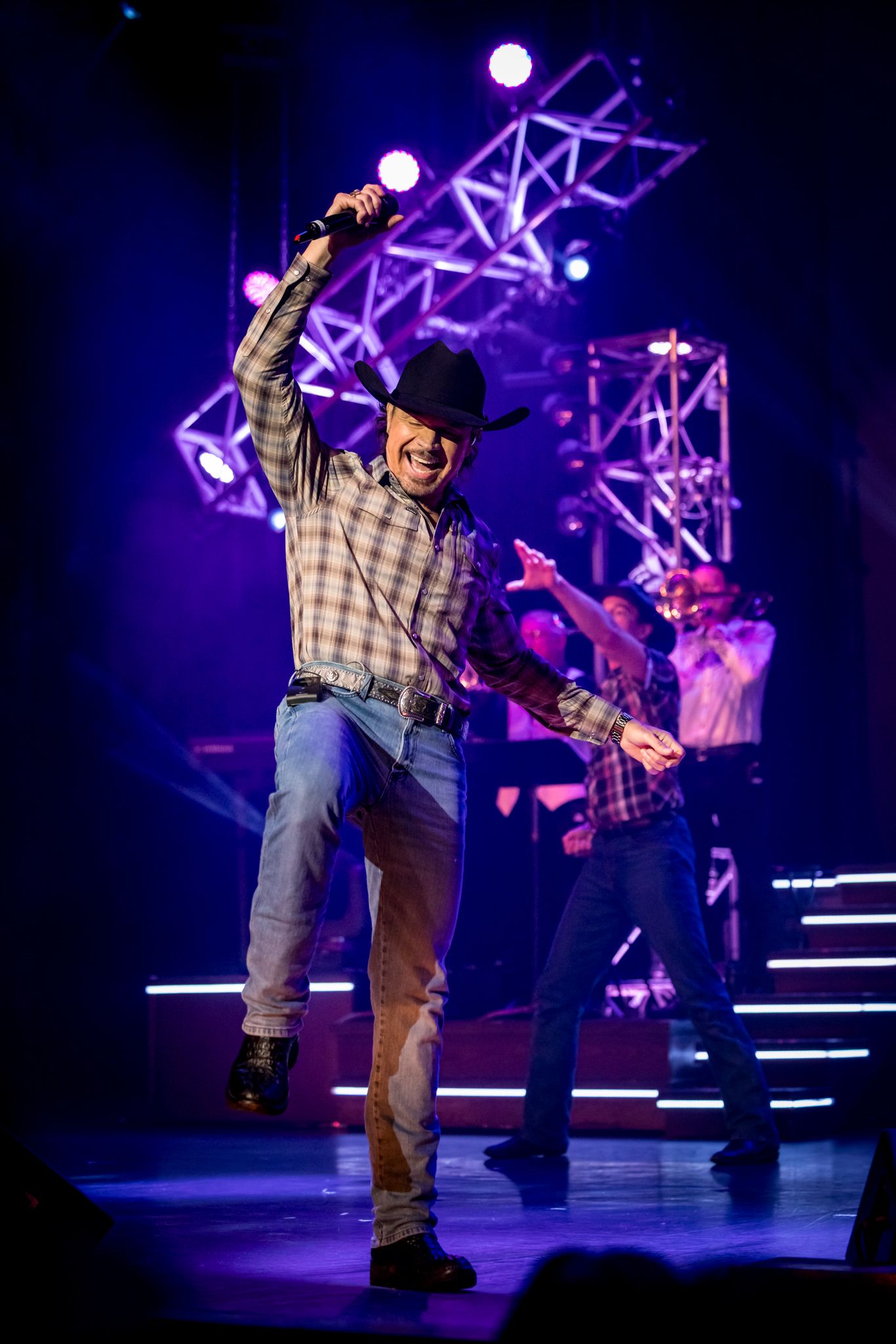 man in jeans and a cowboy hat is holding microphone celebrating with his arm in the air on stage