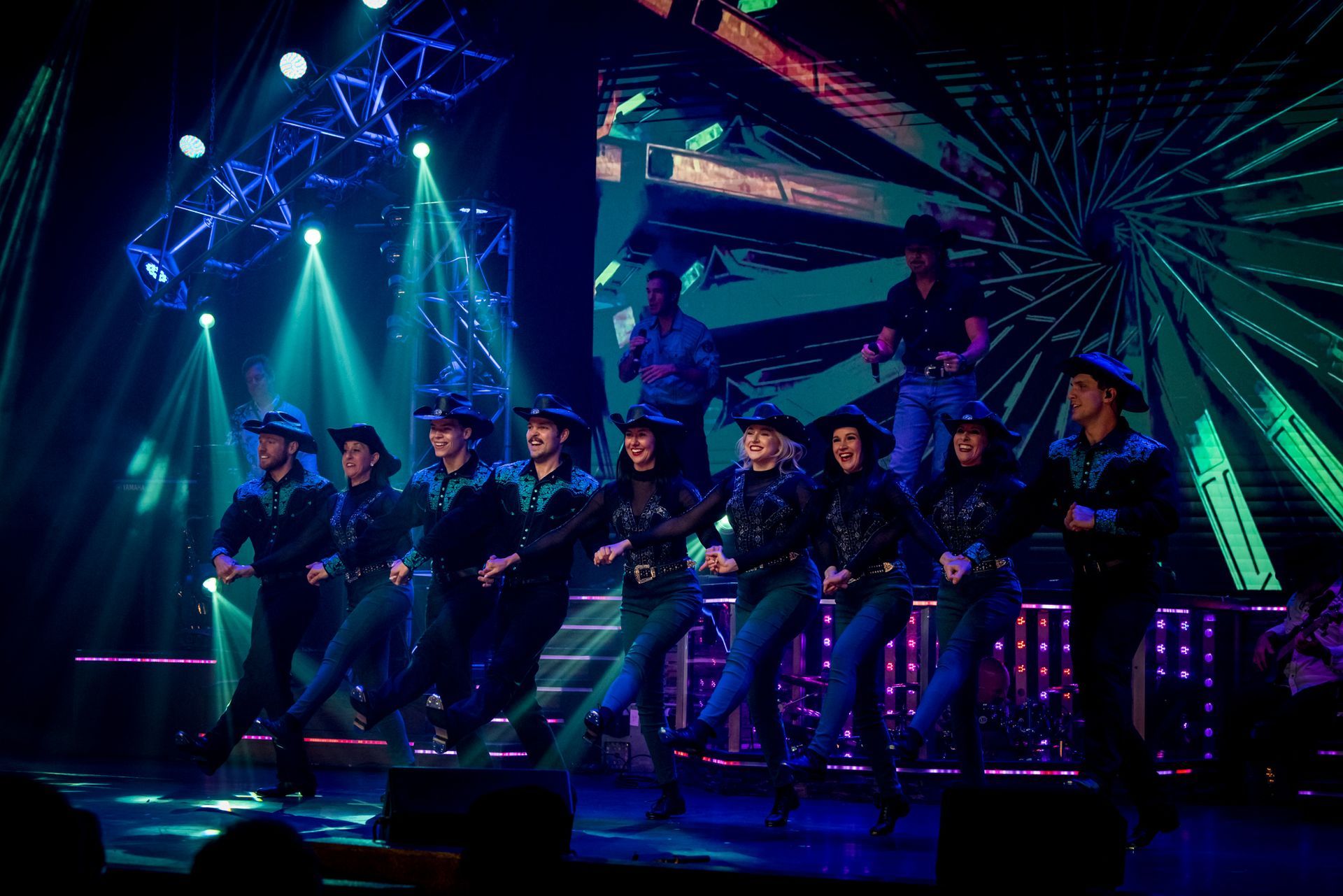 A group of performers in green shirts and black pants dance on a stage under bright spotlights before an audience.