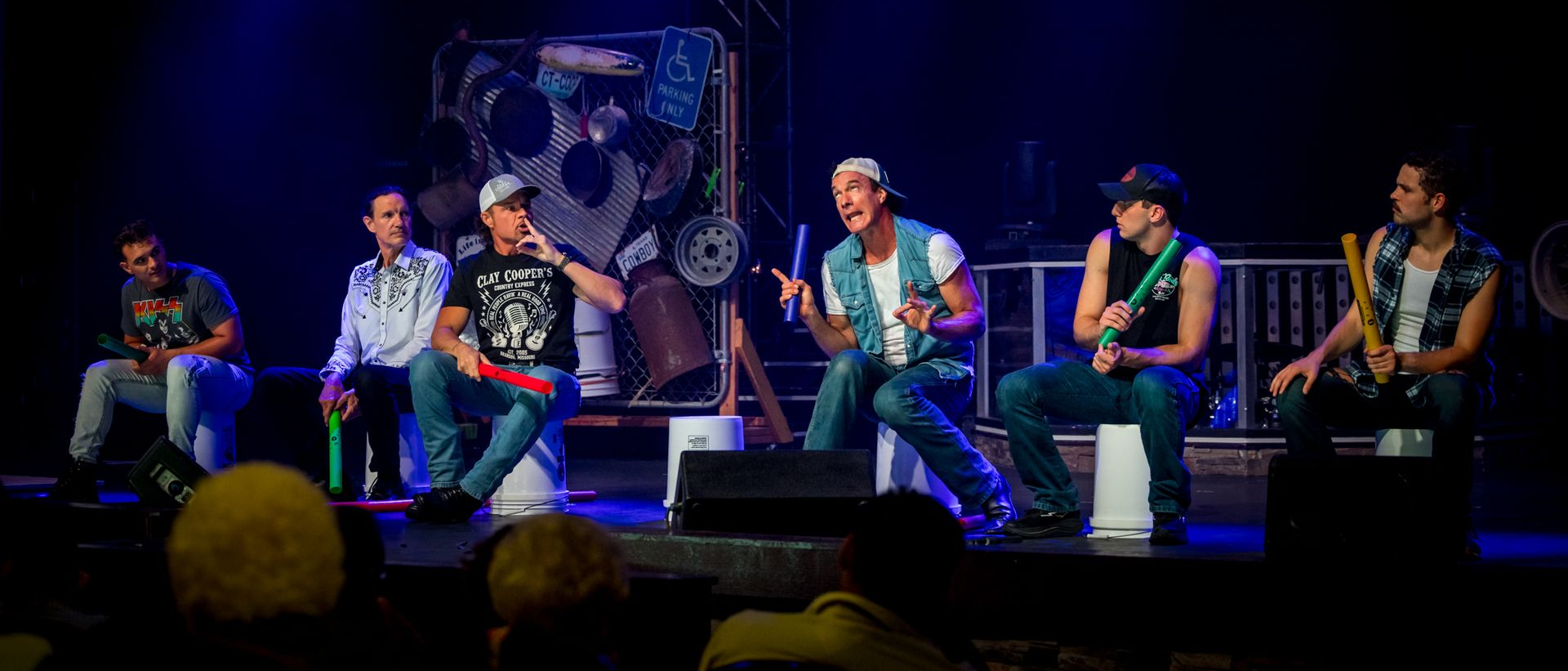 Six people seated on a lit stage, playing hand drums and percussion during a live performance