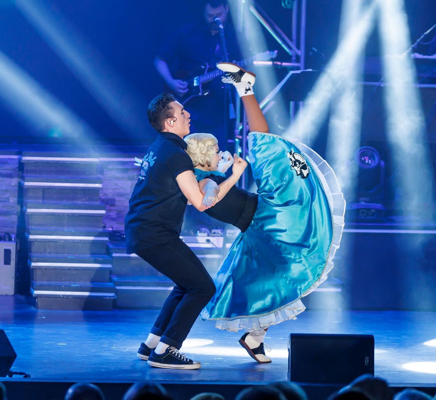 A couple performs a dance on stage; the partner wears a bright blue skirt while kicking their leg high into the air.