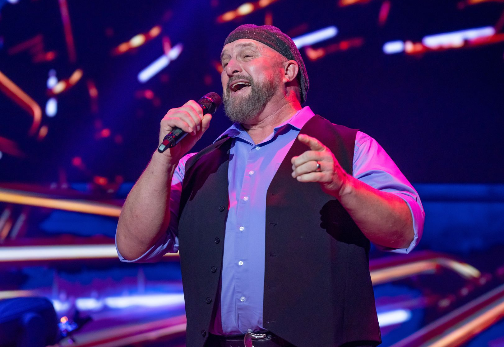 A man in a light blue shirt, dark vest, and knit cap singing into a microphone on a stage with blurred, colorful lights.