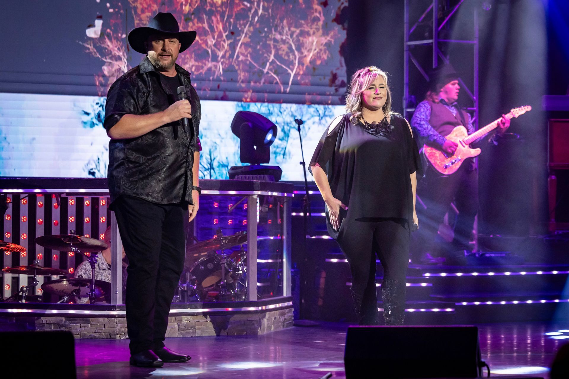 A man in a cowboy hat and a woman stand on a stage with a guitarist behind them, all performing under bright lights.