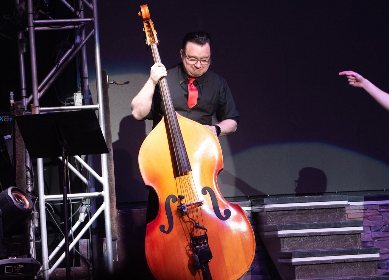 A musician in a black shirt and red tie plays a large, light-colored upright bass on a stage with metal lighting rigs.