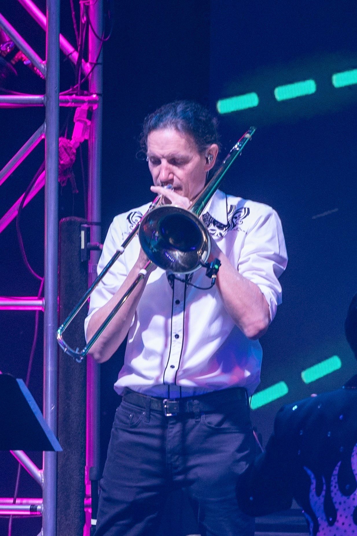 A performer in a white shirt plays the trombone on a stage with purple-lit scaffolding and a digital blue screen.