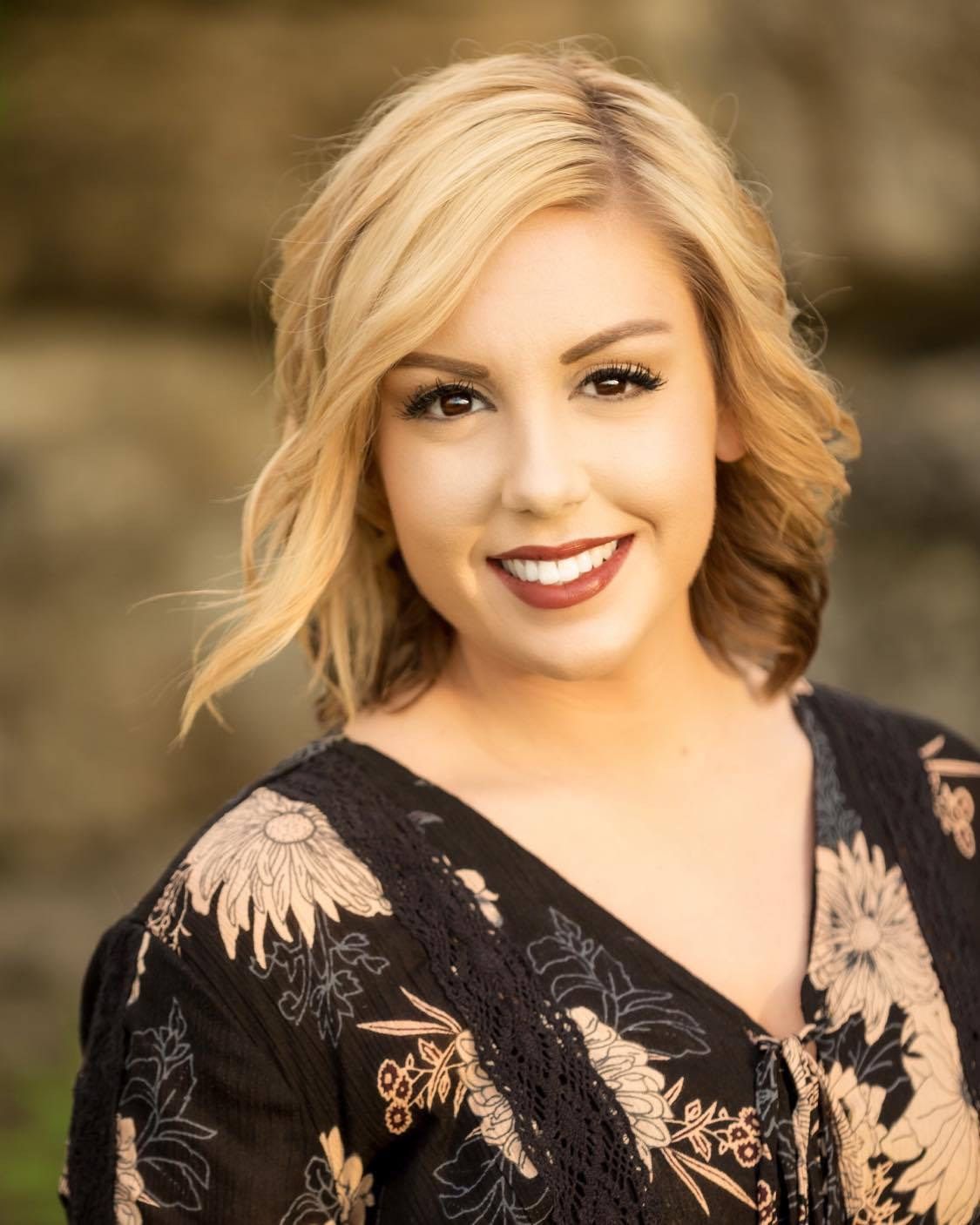 A smiling blonde woman with dark lipstick wears a black floral top, standing against a blurred, light-colored background.