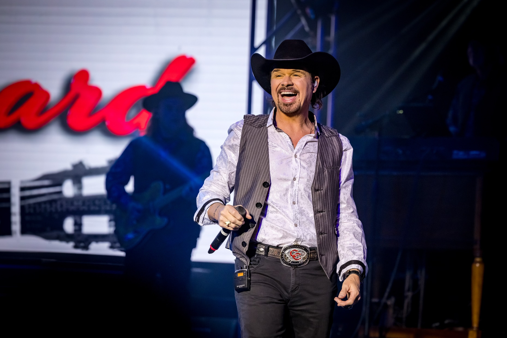 A performer in a cowboy hat and vest sings into a microphone on stage, with a guitar silhouette visible on the screen.