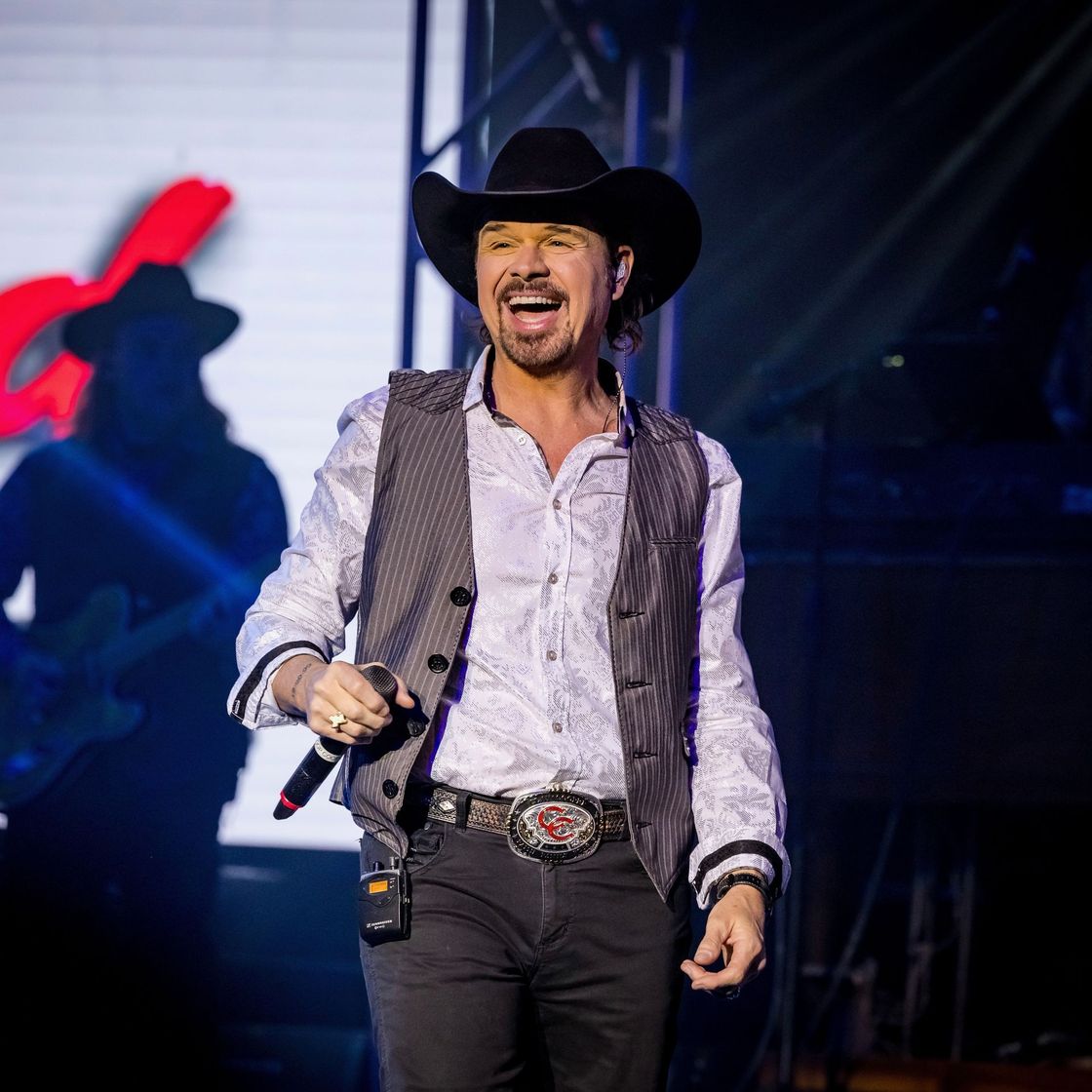 Smiling person wearing a black cowboy hat, silver button-down shirt, and striper vest on stage with a microphone in hand 