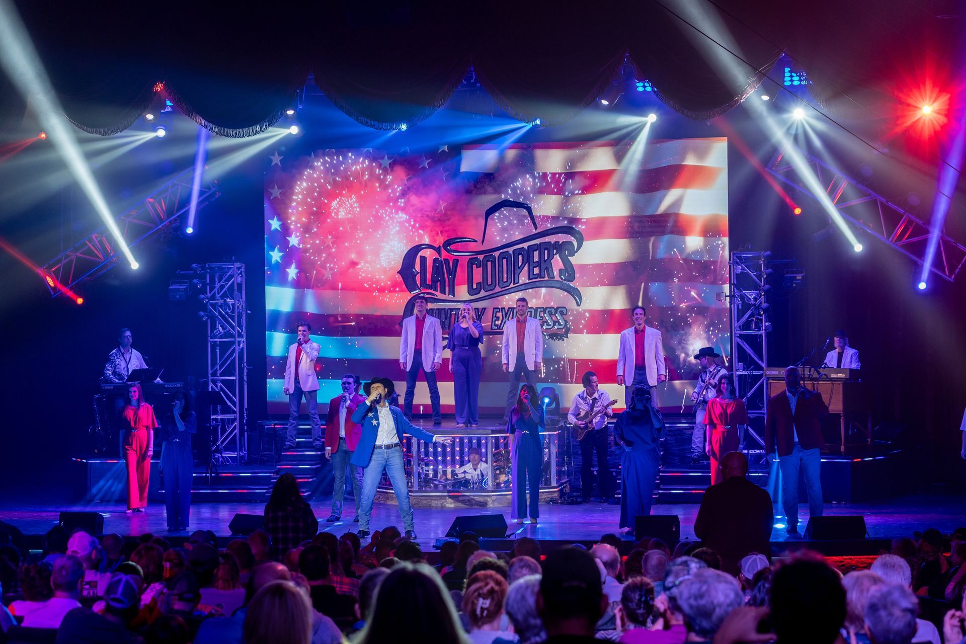 A stage performance with singers and musicians in front of a giant screen displaying an American flag and fireworks.