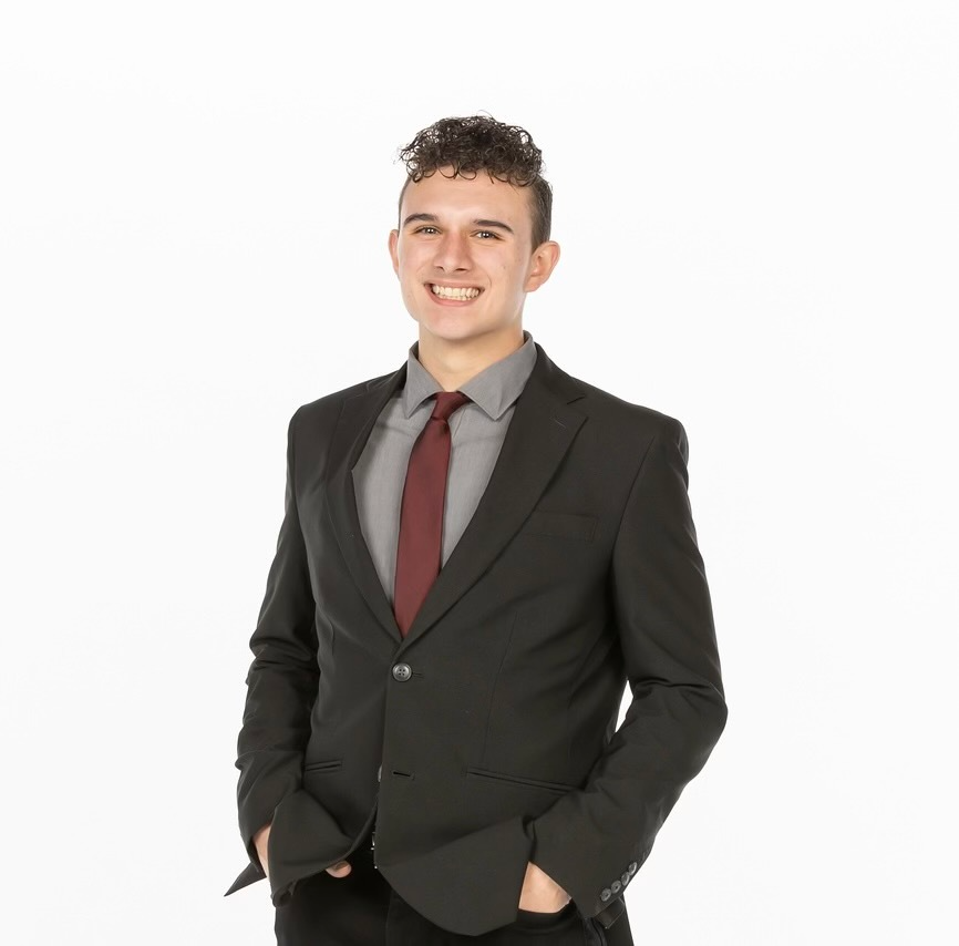 A person smiling in a black suit, grey collared shirt, and maroon tie against a white background with hands in pockets.