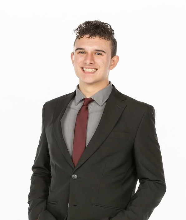 A person in a black suit, grey shirt, and maroon tie smiling against a plain white background.