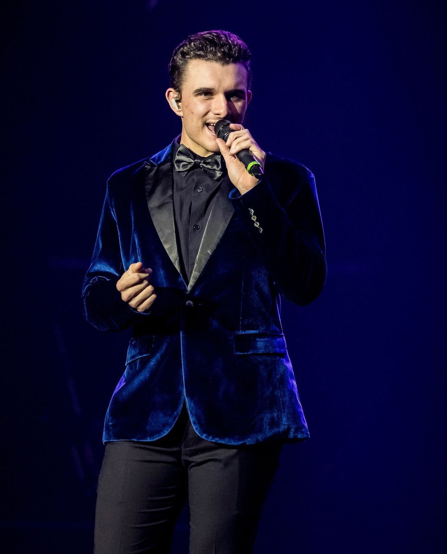 A performer in a blue velvet blazer and bowtie sings into a microphone against a dark blue background.