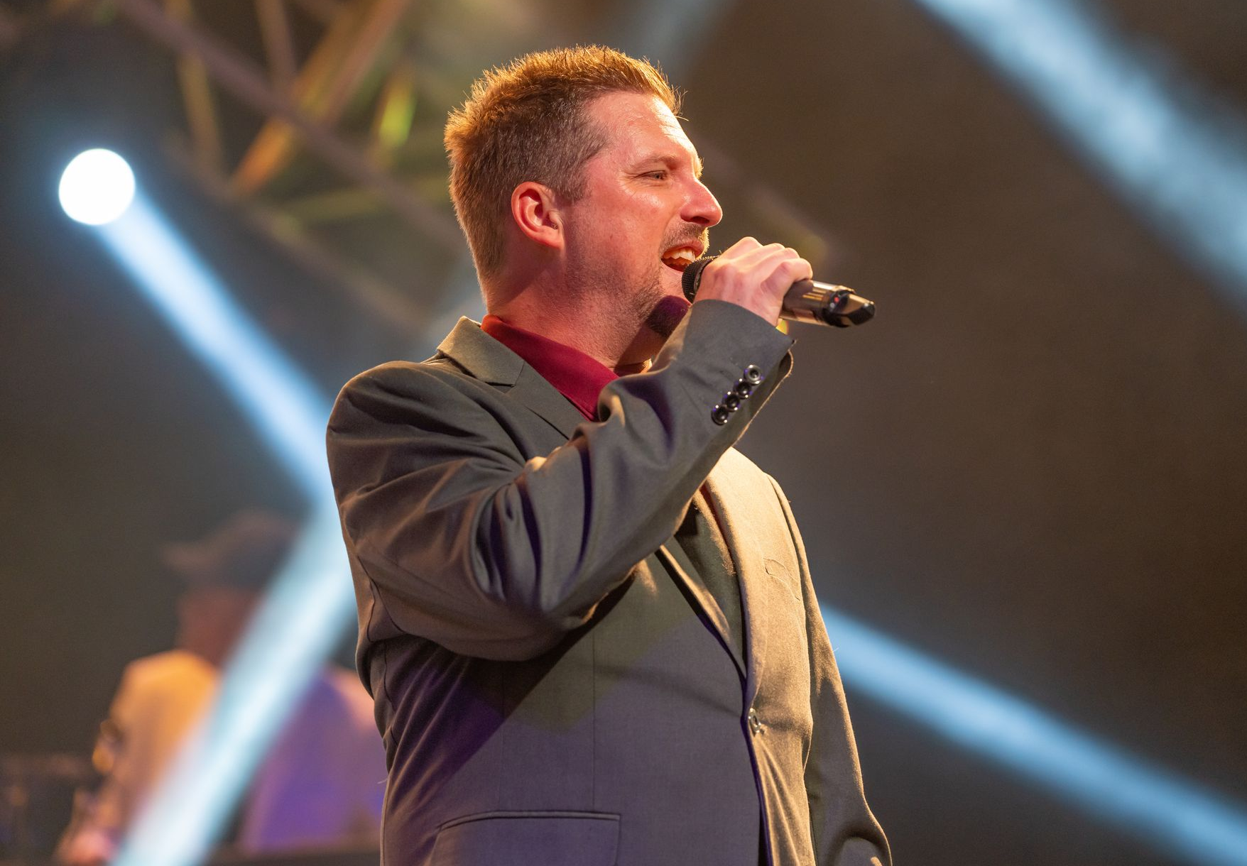A man in a suit sings into a microphone on a stage lit by bright spotlights.