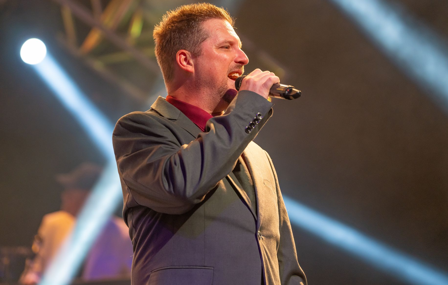 A performer in a grey suit and dark shirt sings into a handheld microphone on a brightly lit stage.
