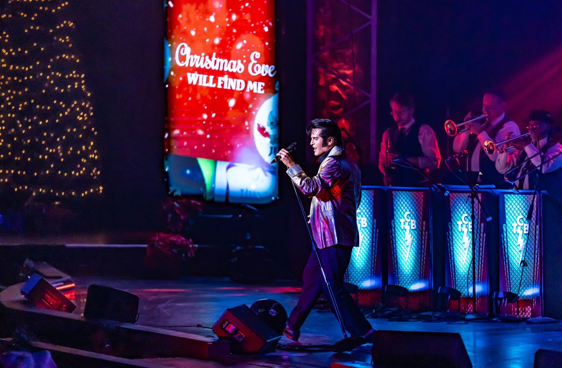 A performer in an Elvis-style suit sings on a stage near a Christmas tree and a band with brass instruments.
